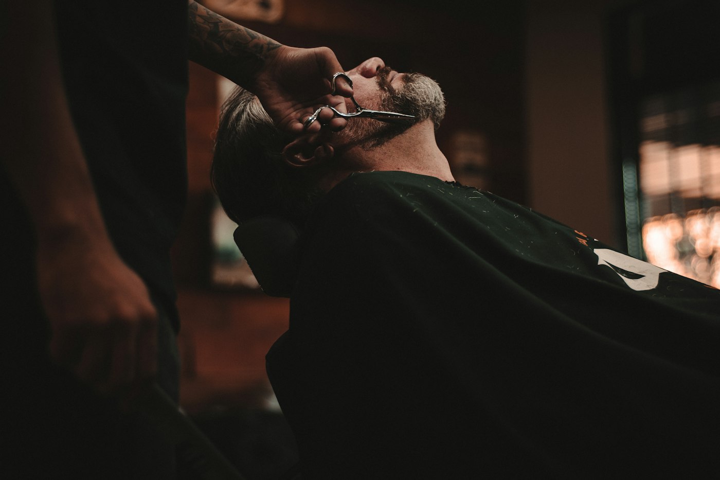 Barber in a dark vintage shop using scissors on a seated guest.