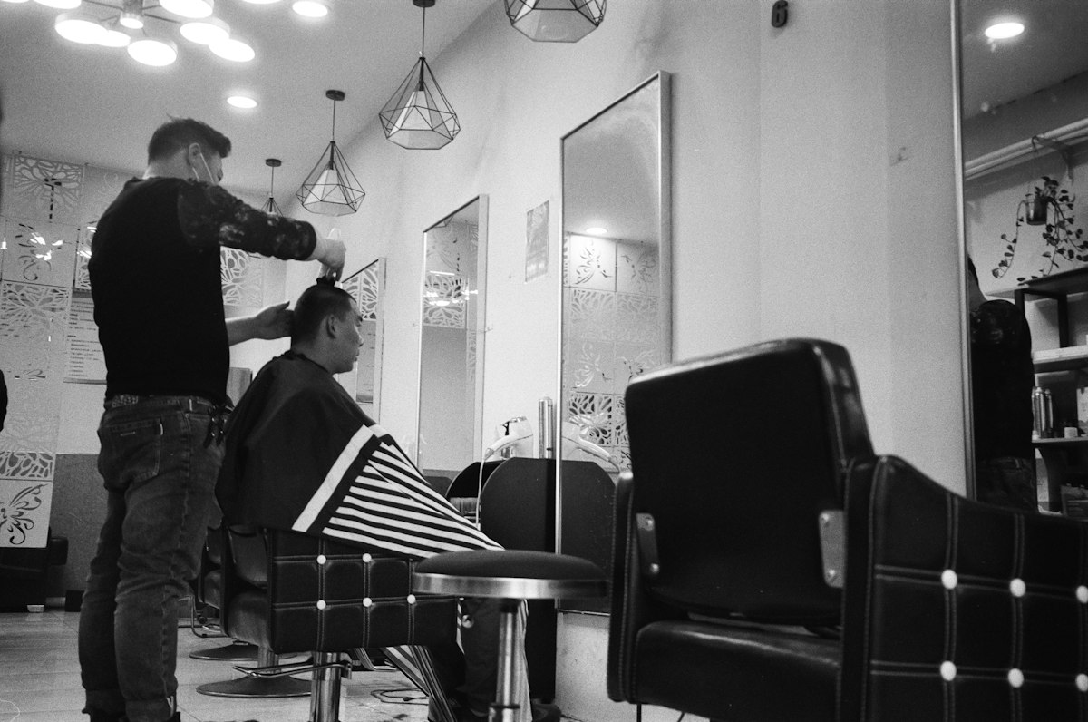 Black-and-white film photograph of a barber cutting a guest’s hair.